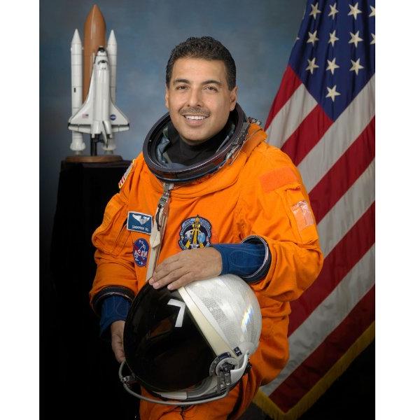 A person wearing an orange jumpsuit and holding an astronaut helmet smiles while sitting. An American flag and a rocket are in the background.